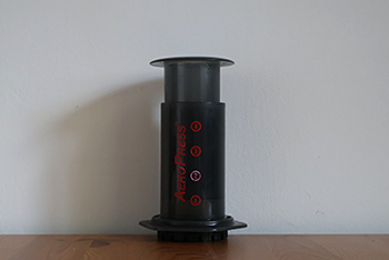 A plastic cylindrical tube with a press on the top and a red 'Aeropress' logo on the side, used to make coffee.