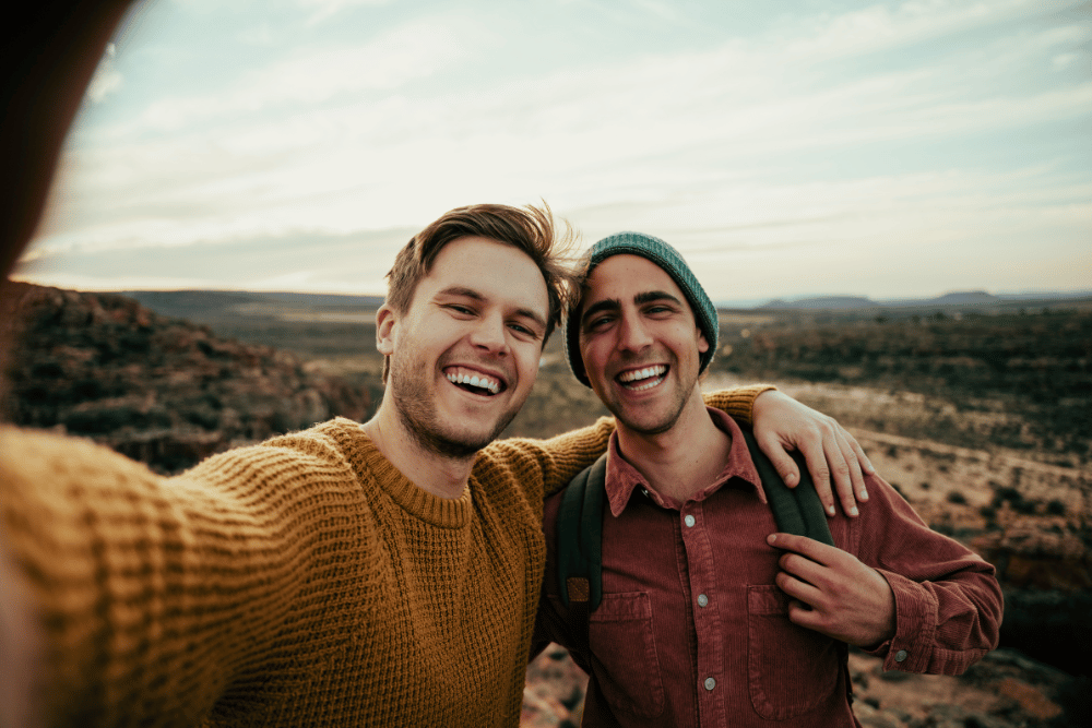 Two male friends smiling at the camera from the top of a mountain at sunset